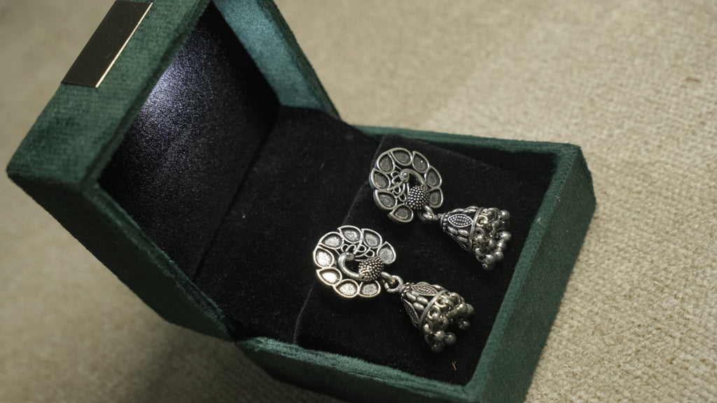 Oxidised sterling silver peacock jhumka earrings presented in a green velvet jewellery box.