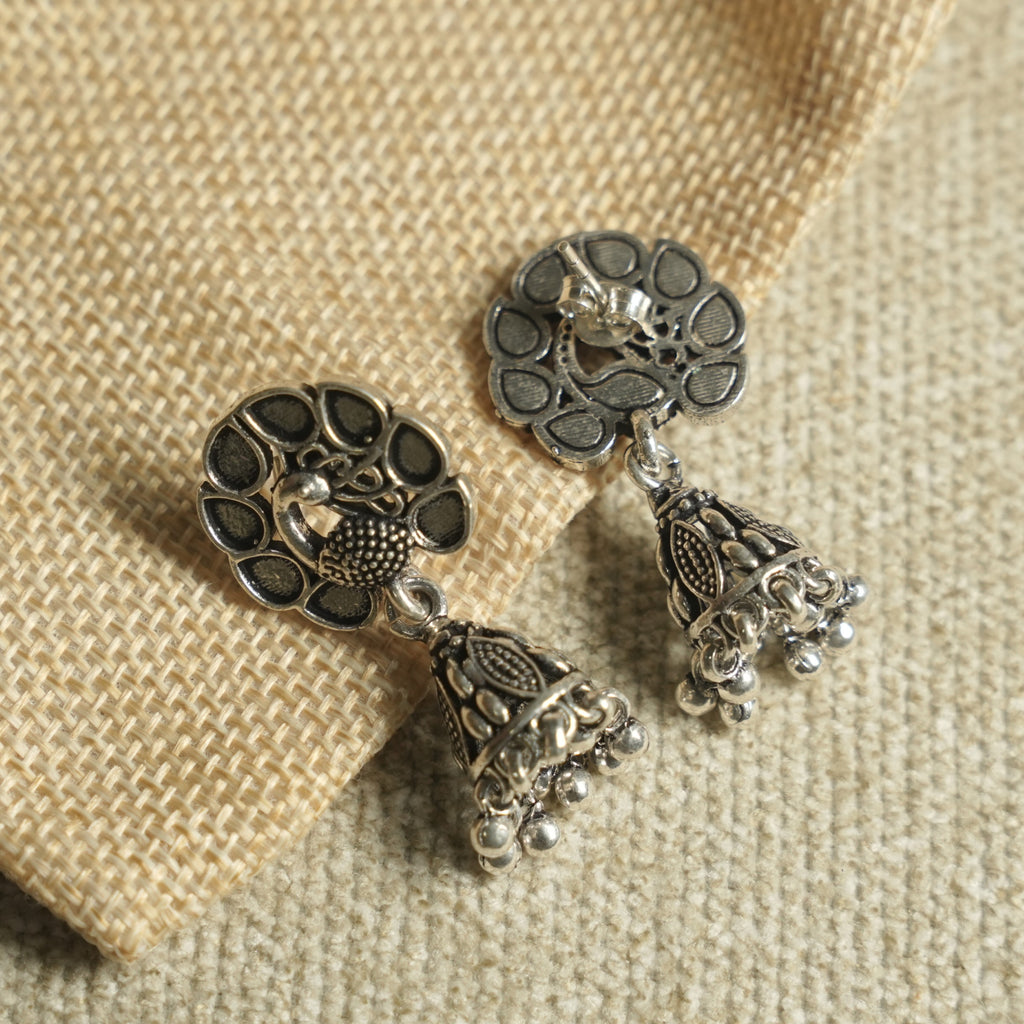 Angled view of the peacock jhumka earrings on jute fabric, showing the push-back post and the fringed bell drops.