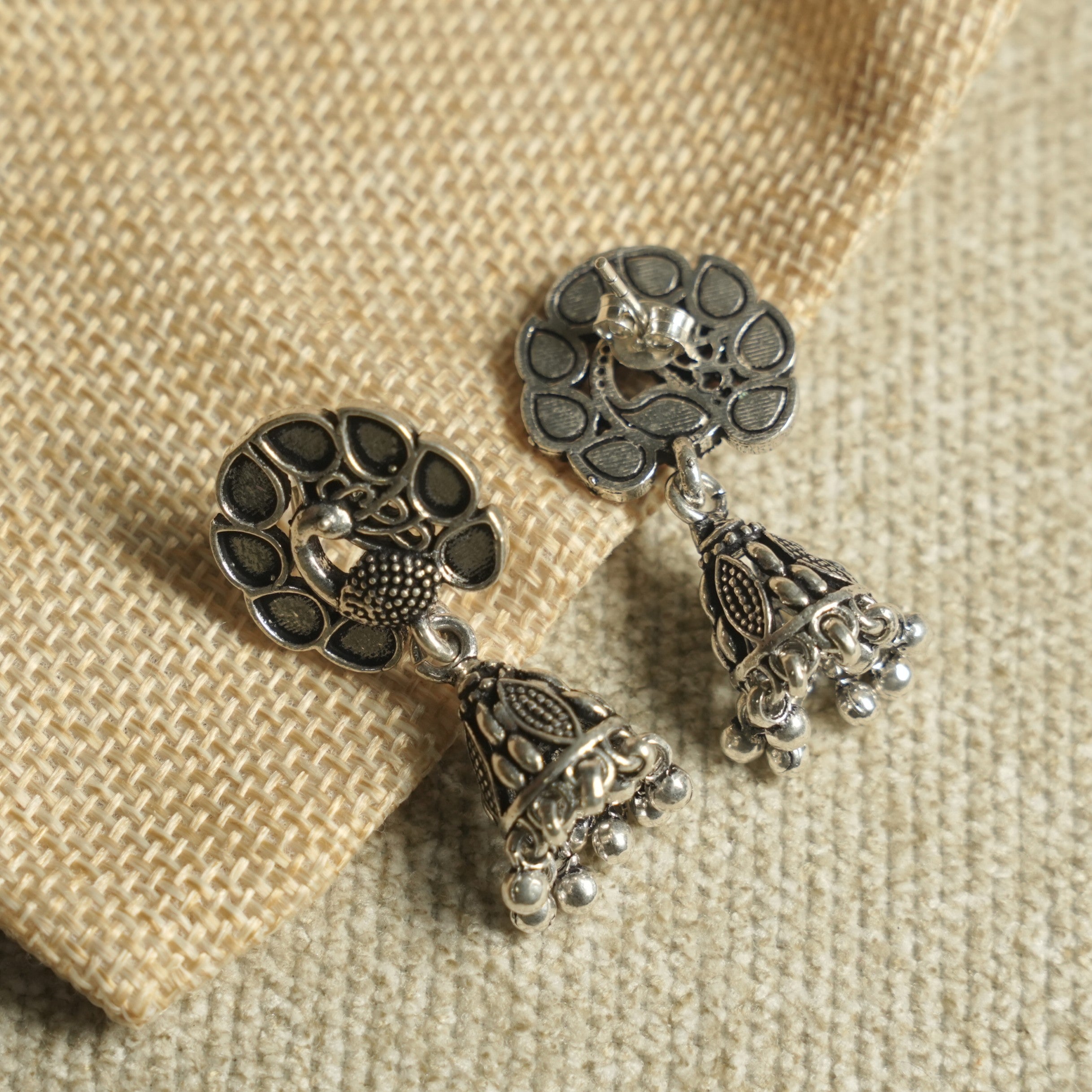 Angled view of the peacock jhumka earrings on jute fabric, showing the push-back post and the fringed bell drops.