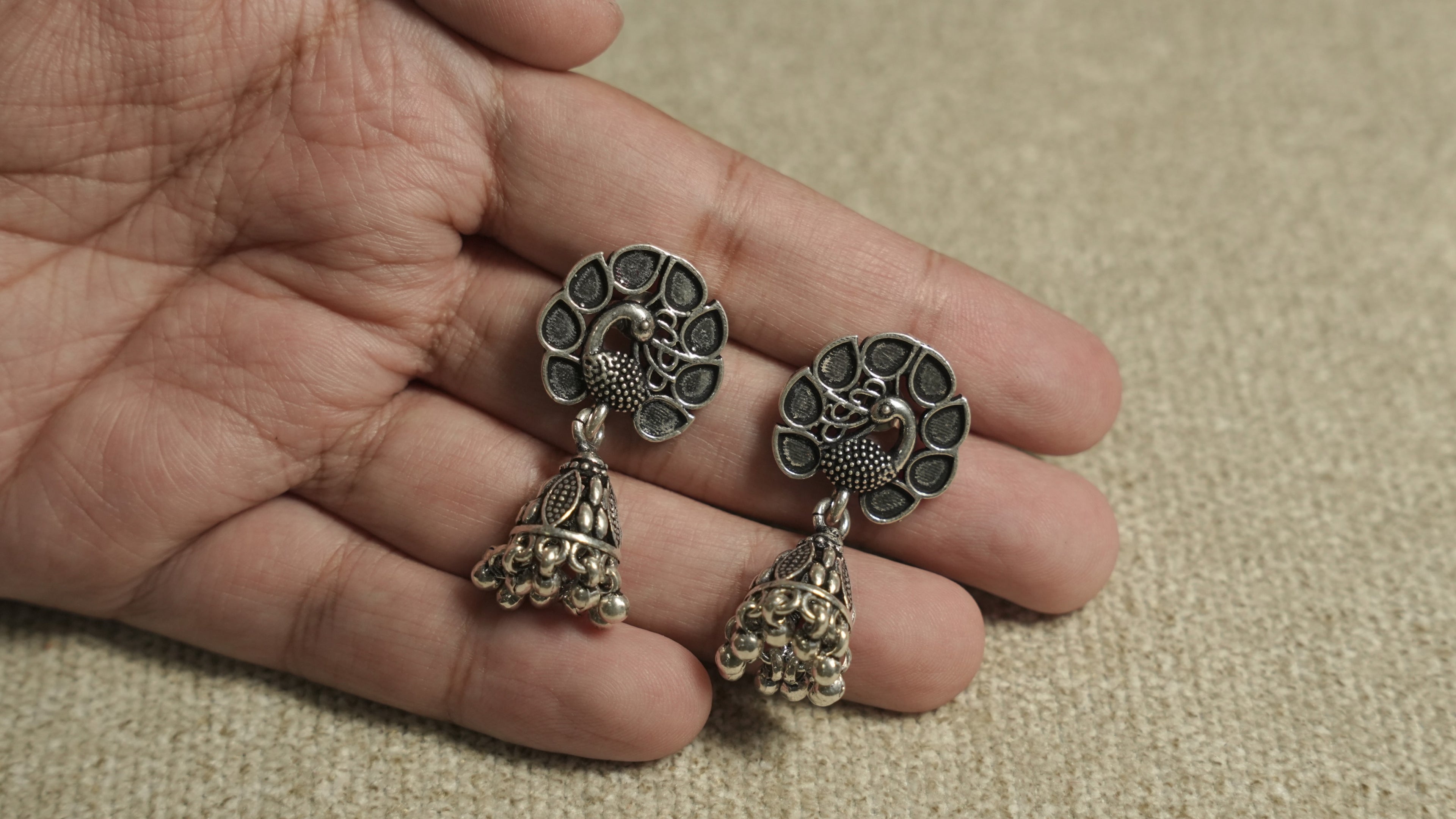 Peacock jhumka earrings held in an open palm, showing their compact everyday scale.
