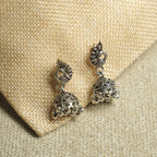 Pair of oxidised sterling silver peacock jhumka earrings with domed, bead-fringed bells, resting on jute fabric.