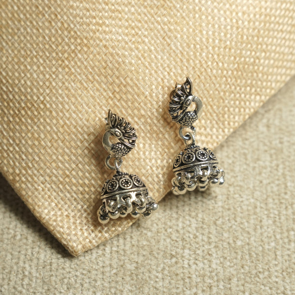 Pair of oxidised sterling silver peacock jhumka earrings with domed, bead-fringed bells, resting on jute fabric.