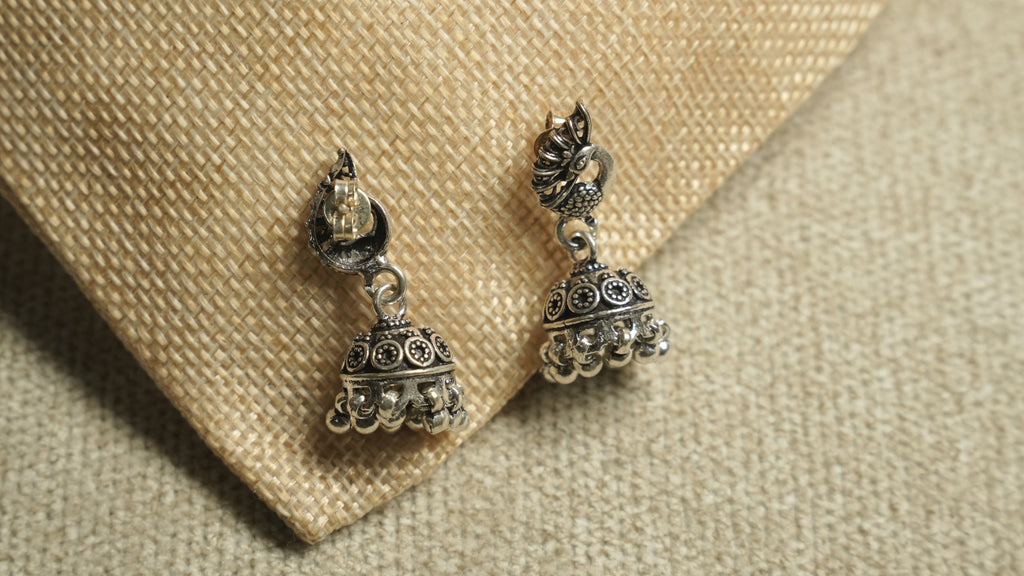 Front and back view of the peacock jhumka earrings on jute, showing the post fitting and the patterned bell drops.