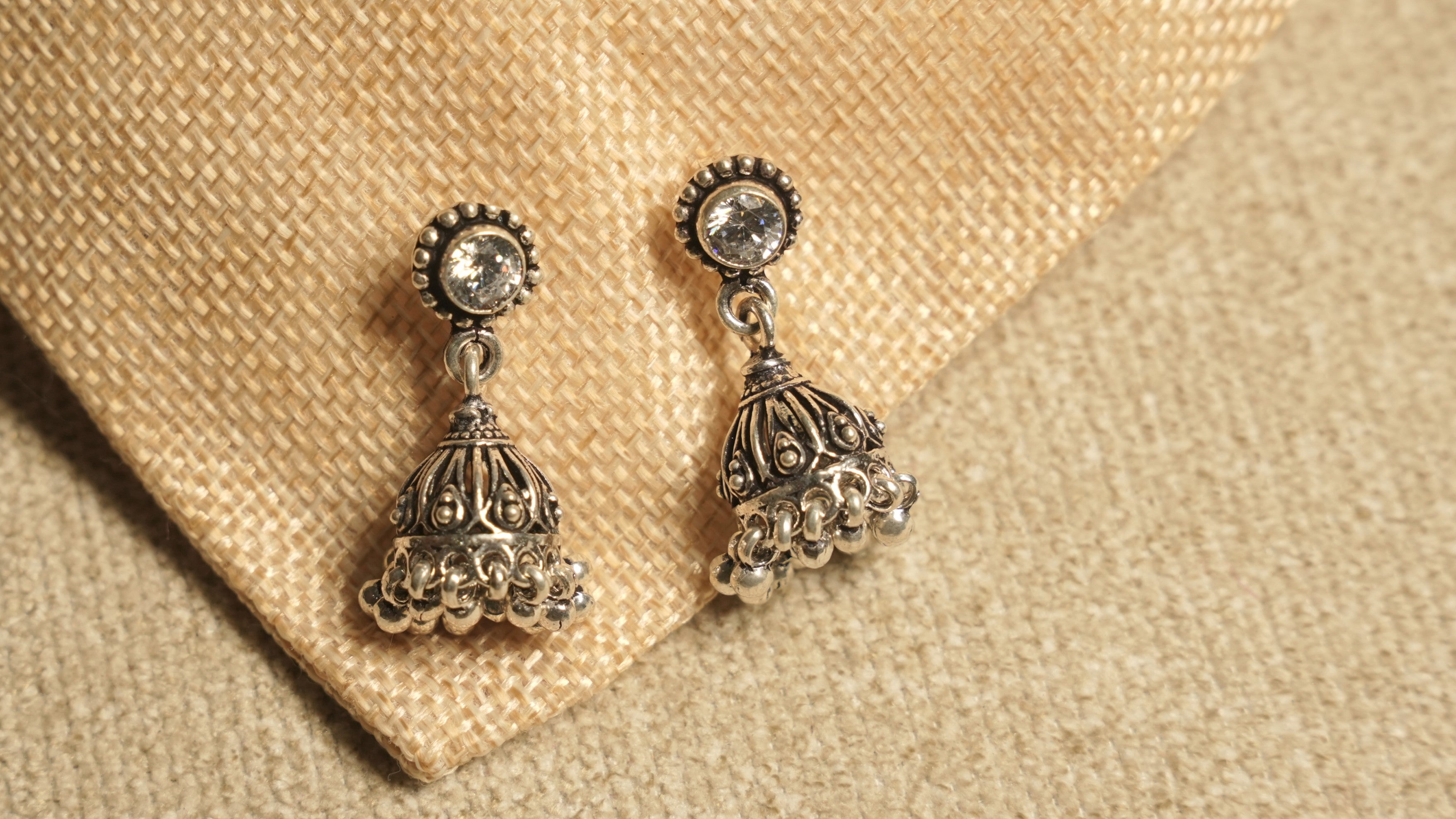 Oxidised sterling silver jhumka earrings topped with round cubic zirconia studs, resting on jute fabric.