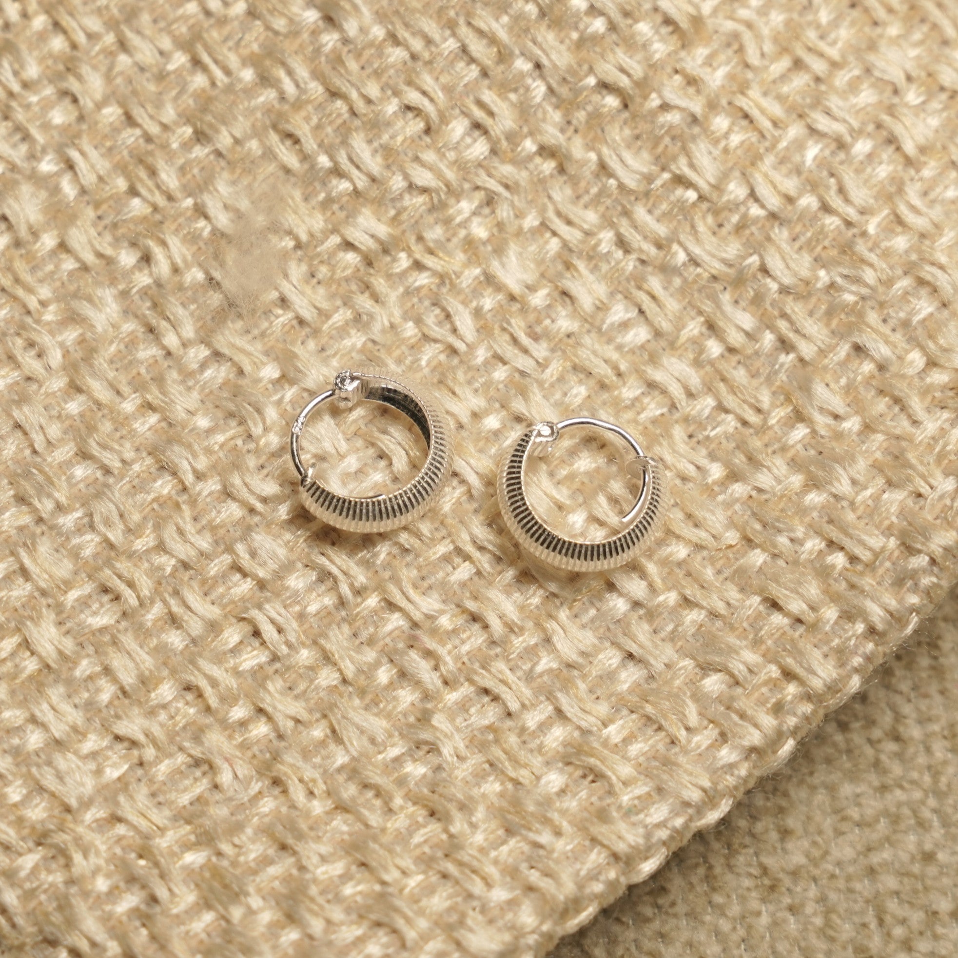 Pair of small sterling silver huggie hoops with ribbed texture, resting on a woven pouch with gold drawstring.