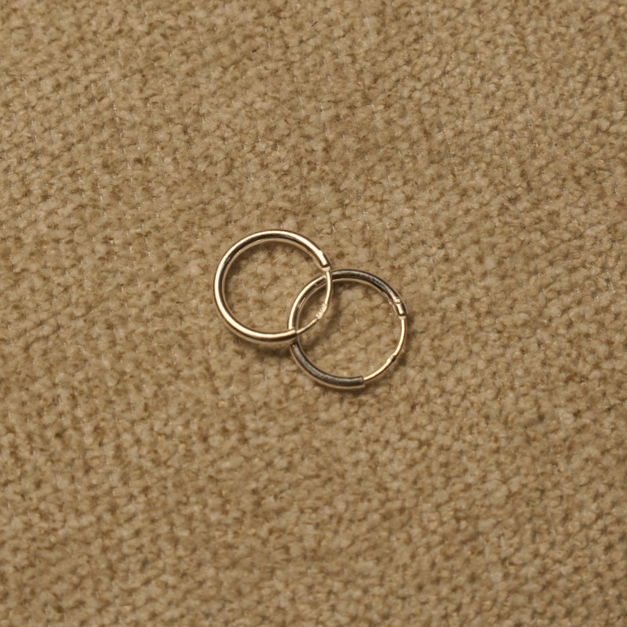 Pair of slim polished sterling silver hoop earrings overlapping on beige fabric.
