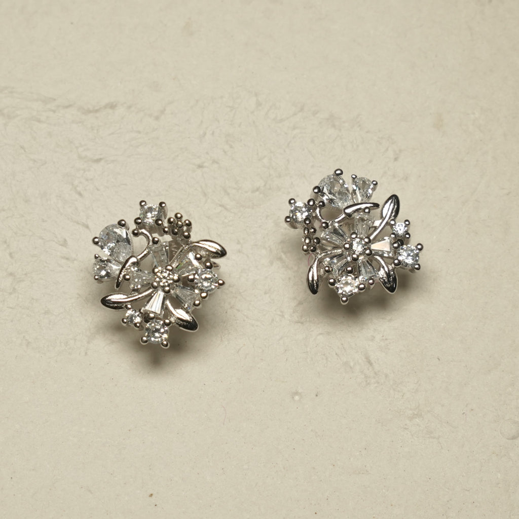 Pair of sterling silver floral cluster stud earrings with pear, baguette and round cubic zirconia set among polished silver leaves, on a pale stone backdrop.