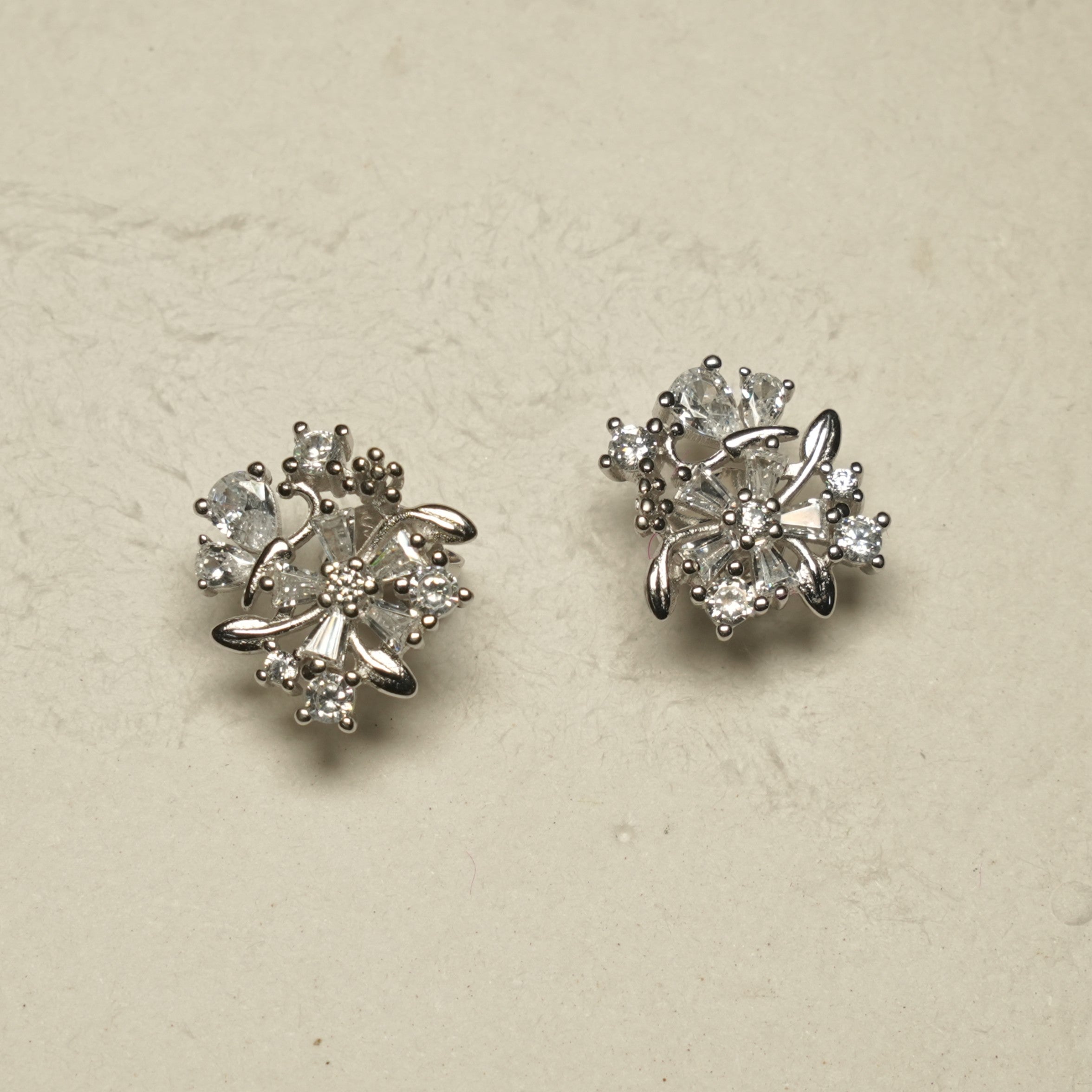Pair of sterling silver floral cluster stud earrings with pear, baguette and round cubic zirconia set among polished silver leaves, on a pale stone backdrop.