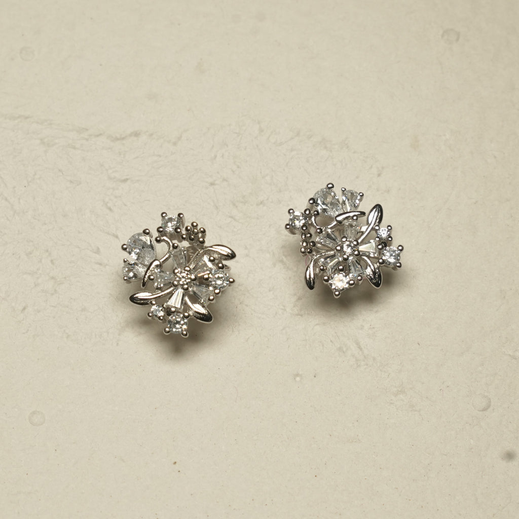 Angled view of the sterling silver floral stud earrings showing the layered cubic zirconia petals and leaf details against pale textured stone.