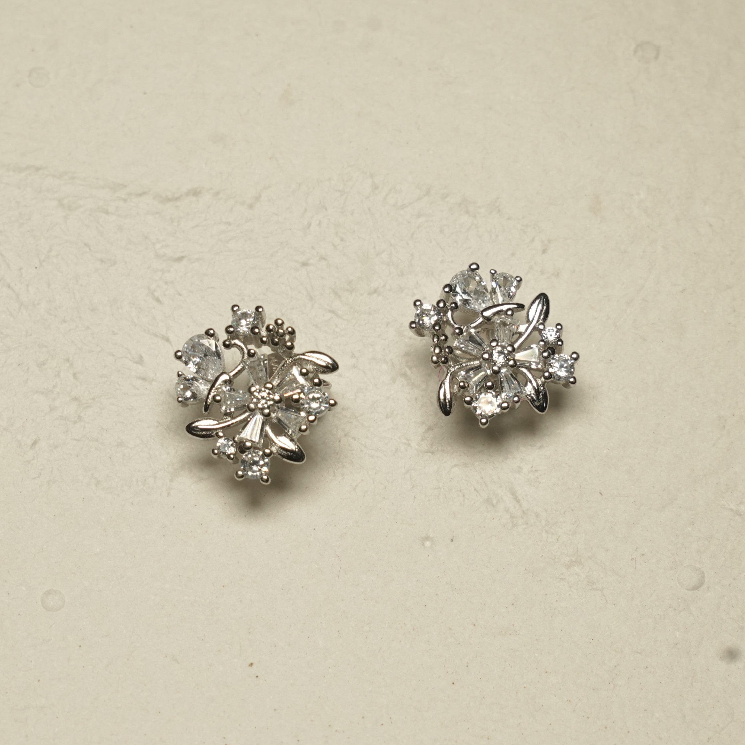 Angled view of the sterling silver floral stud earrings showing the layered cubic zirconia petals and leaf details against pale textured stone.