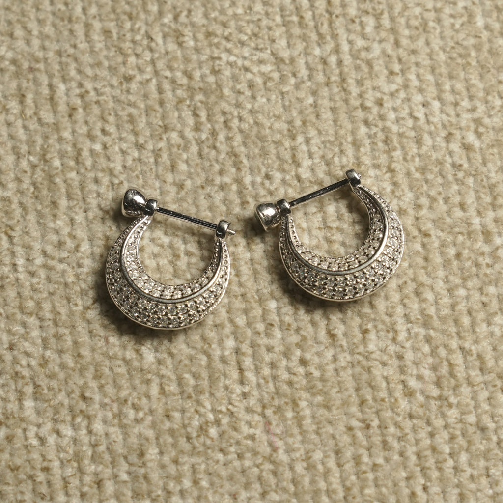 Pair of sterling silver crescent hoop earrings paved with rows of small cubic zirconia, each hanging from a bar-and-bead closure, on beige woven fabric.