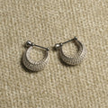 Pair of sterling silver crescent hoop earrings paved with rows of small cubic zirconia, each hanging from a bar-and-bead closure, on beige woven fabric.