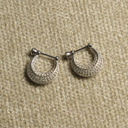 Pair of sterling silver crescent hoop earrings paved with rows of small cubic zirconia, each hanging from a bar-and-bead closure, on beige woven fabric.