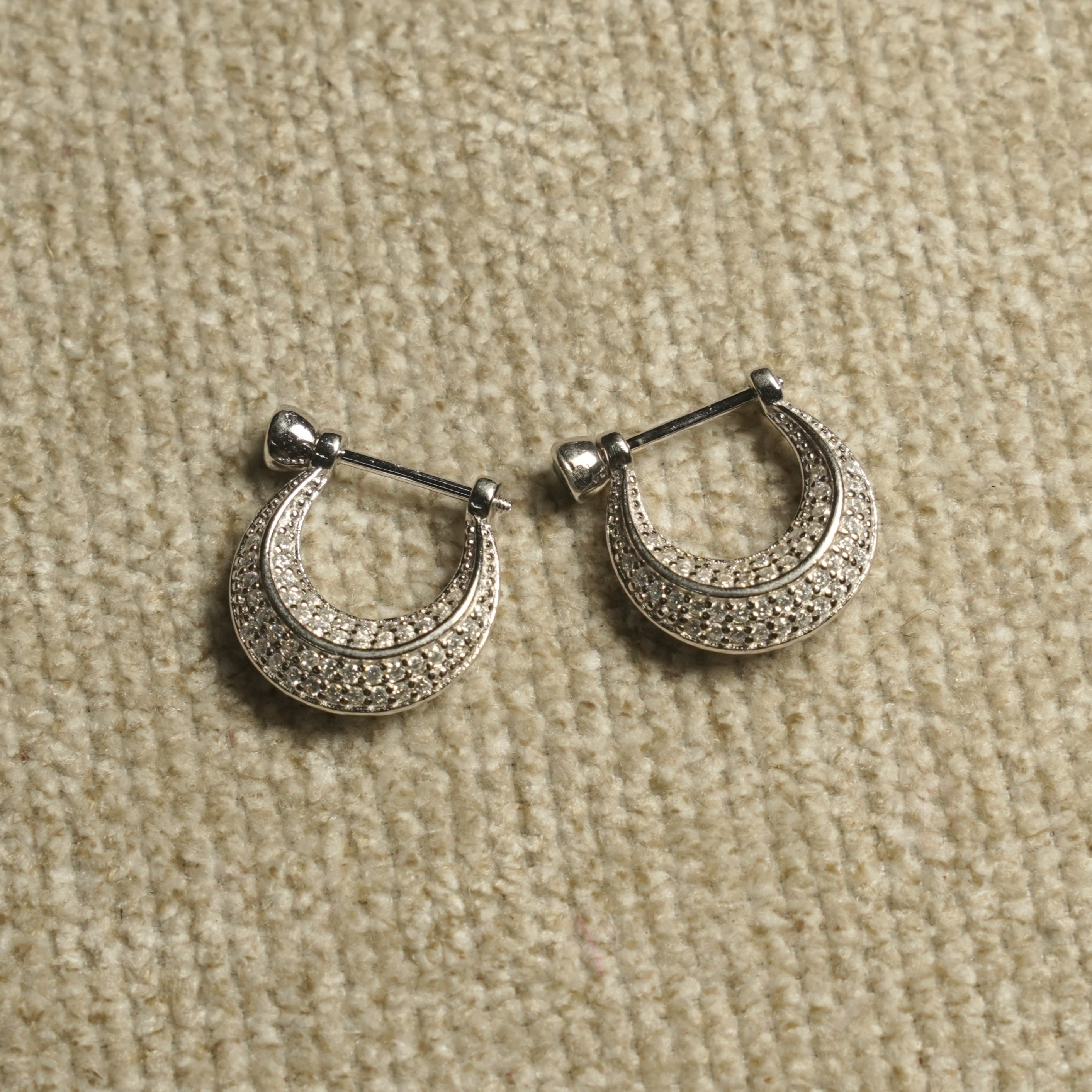 Pair of sterling silver crescent hoop earrings paved with rows of small cubic zirconia, each hanging from a bar-and-bead closure, on beige woven fabric.