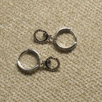 Pair of sterling silver hinged hoop earrings, each with a small bead-textured open-circle charm holding a tiny leaf detail, on beige woven fabric.