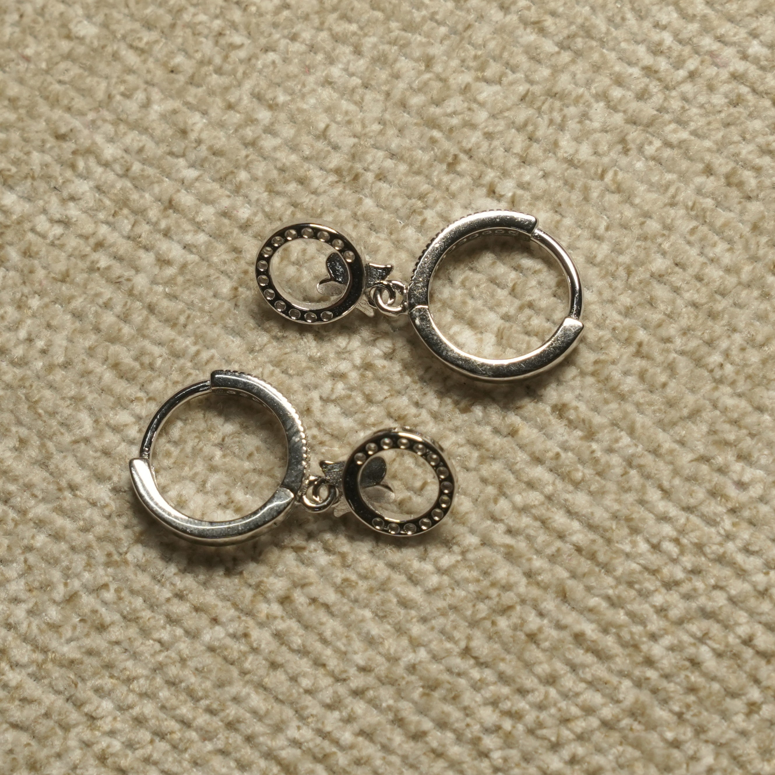 Pair of sterling silver hinged hoop earrings, each with a small bead-textured open-circle charm holding a tiny leaf detail, on beige woven fabric.