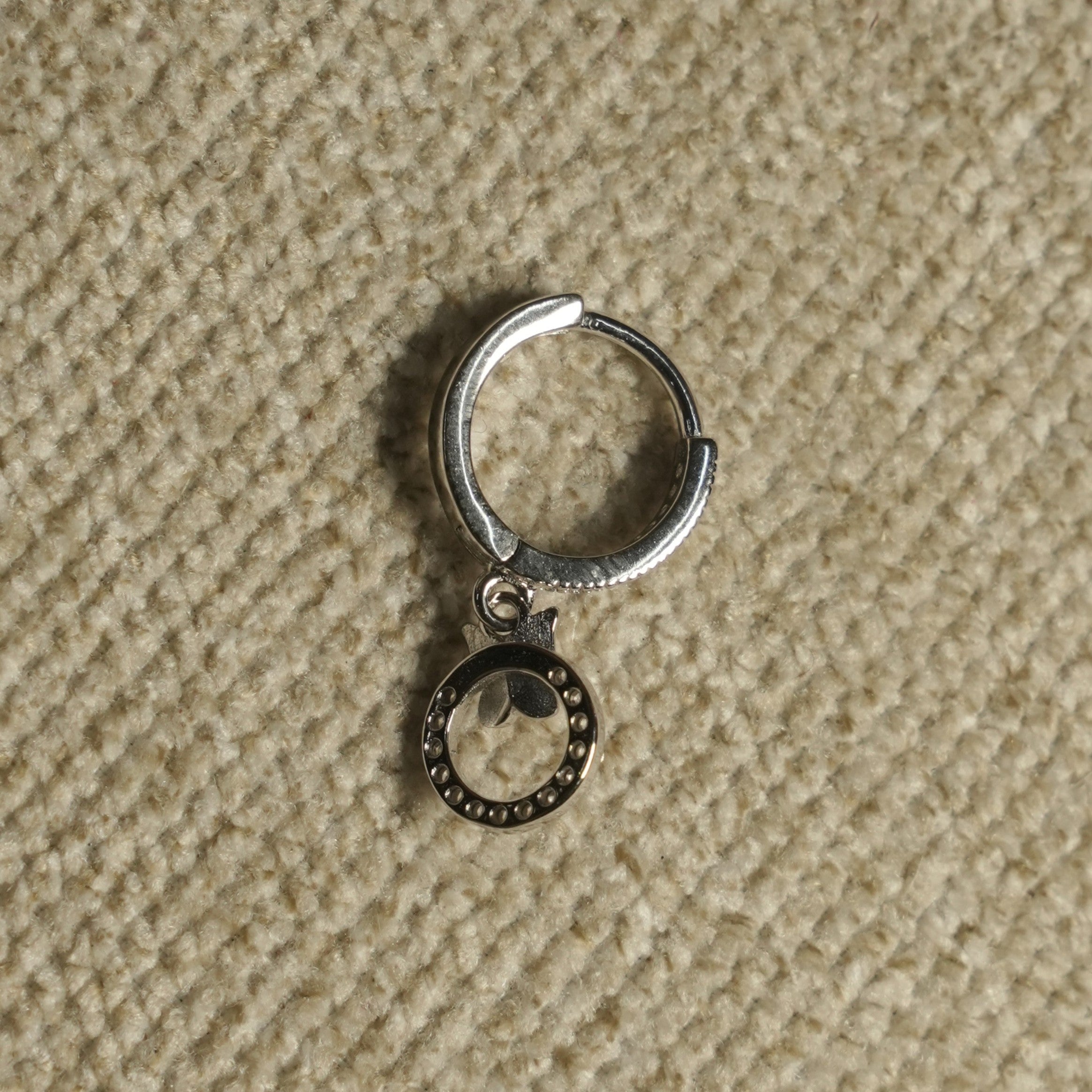 Close view of a single sterling silver hoop earring with its beaded open-circle drop and leaf detail, on beige woven fabric.