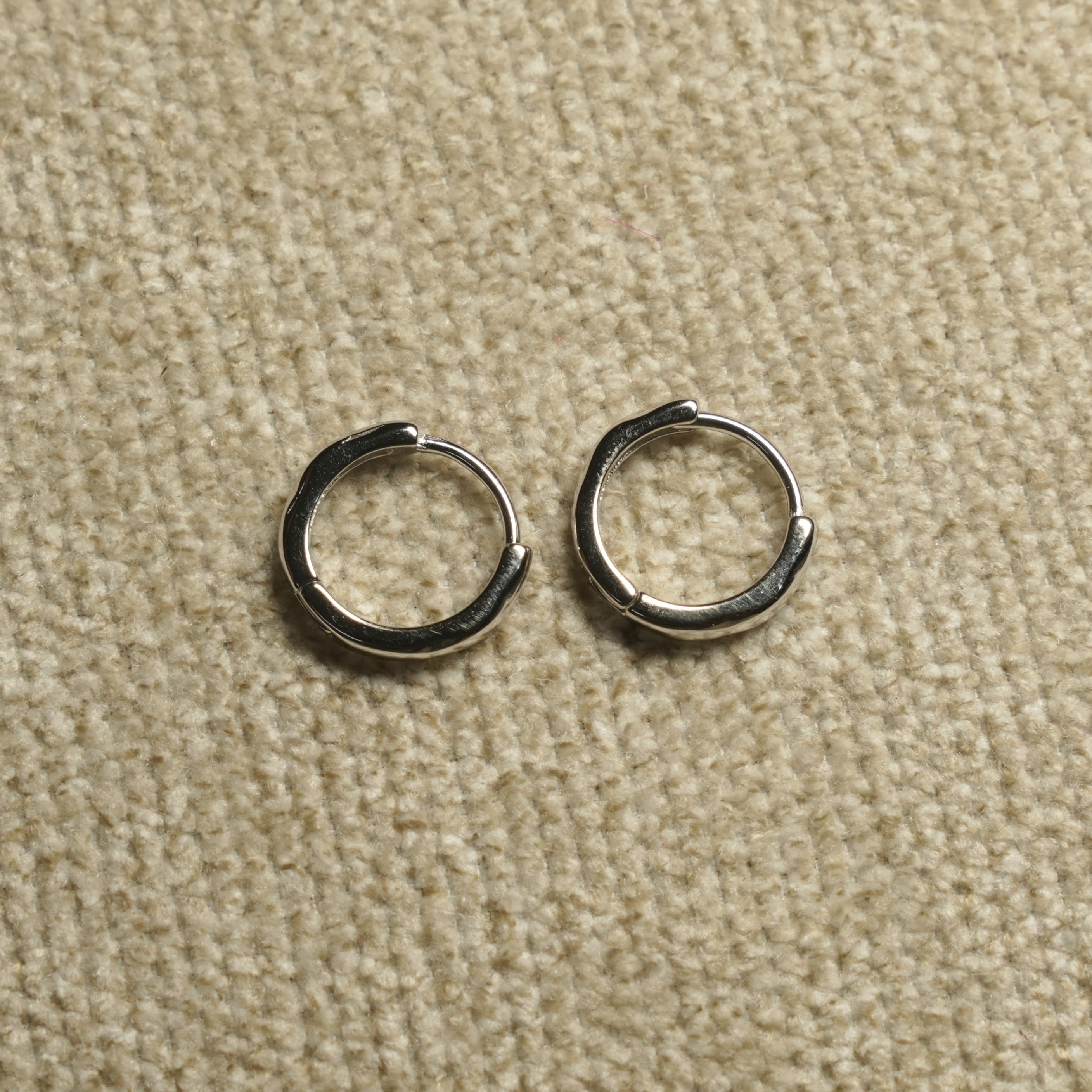 Pair of slim sterling silver huggie hoop earrings with hinged closures, laid side by side on beige woven fabric.