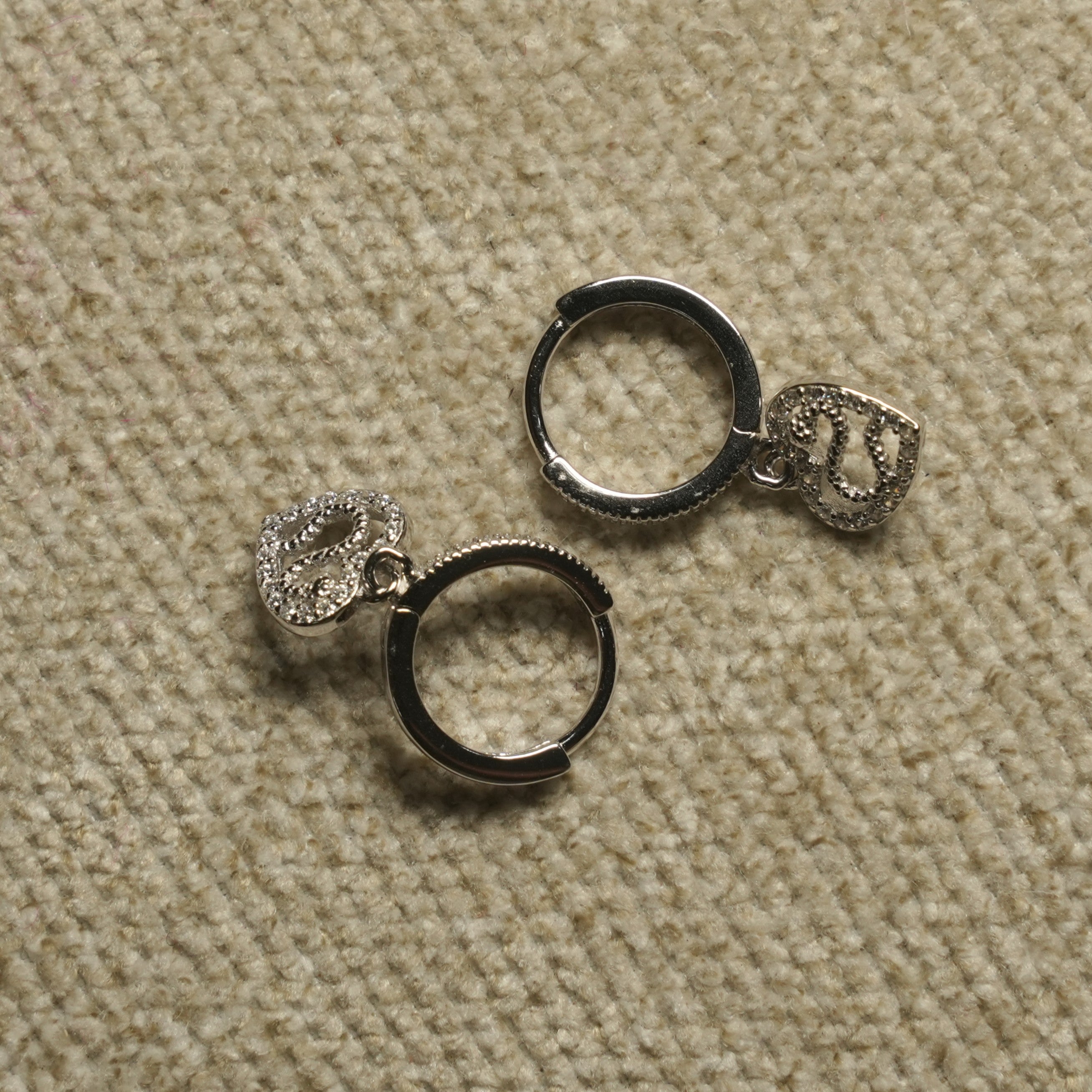Pair of sterling silver hinged hoop earrings, each with a dangling heart charm textured with tiny cubic zirconia, on beige woven fabric.