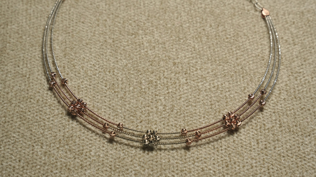 Close view of a three-strand sterling silver necklace with rose gold-tone wire sections and clusters of faceted beads, on beige woven fabric.