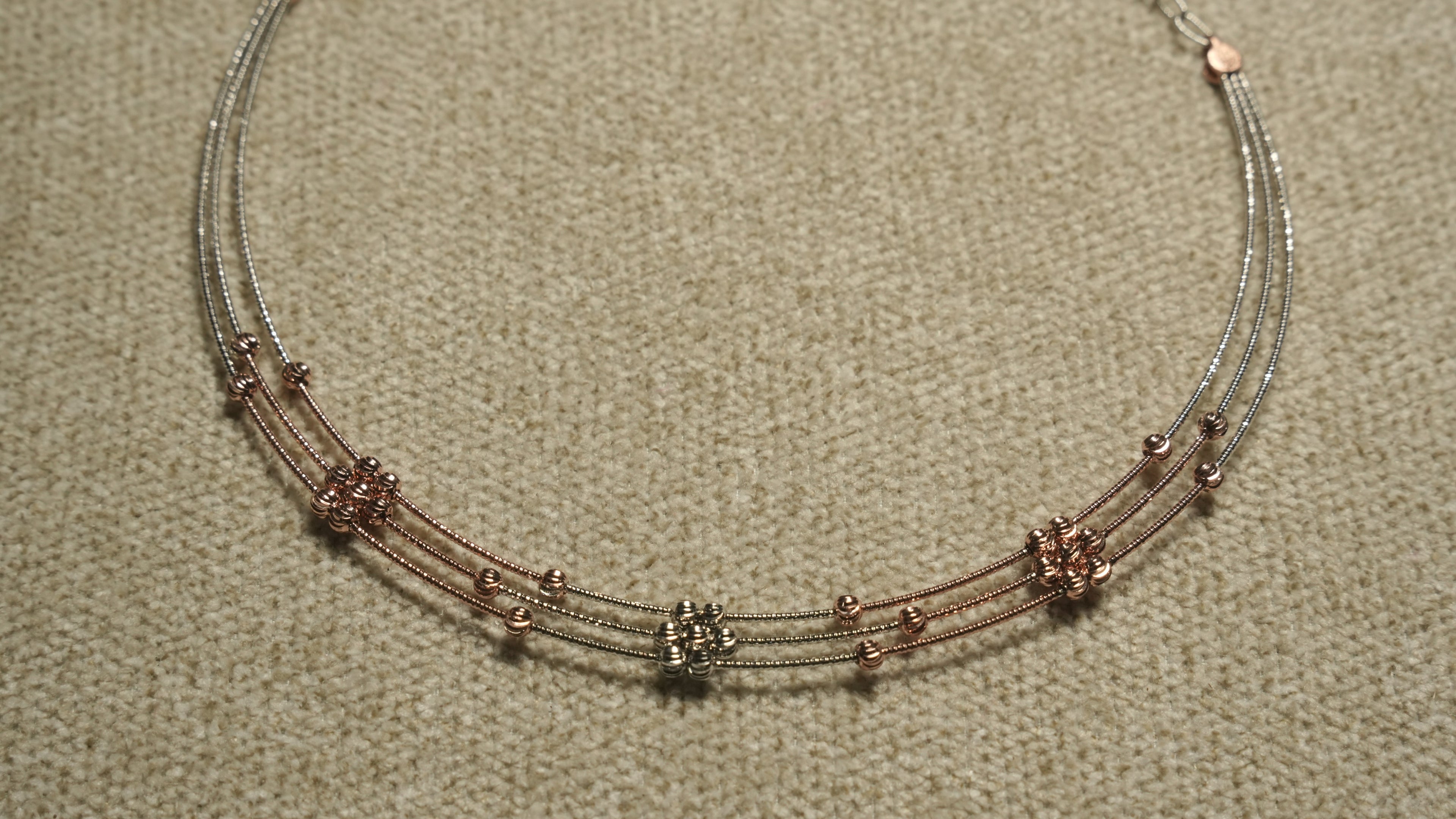 Close view of a three-strand sterling silver necklace with rose gold-tone wire sections and clusters of faceted beads, on beige woven fabric.
