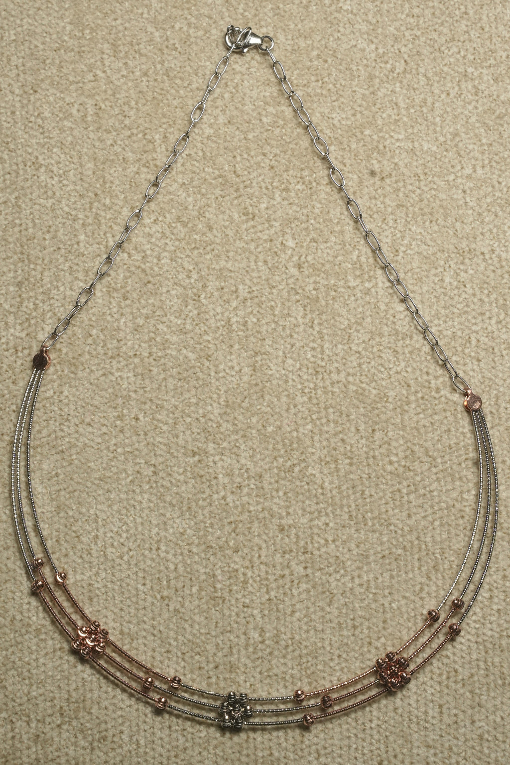 Front section of the three-strand necklace showing rose gold-tone bead clusters and a silver bead cluster at the centre, on beige woven fabric.