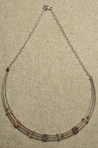 Front section of the three-strand necklace showing rose gold-tone bead clusters and a silver bead cluster at the centre, on beige woven fabric.