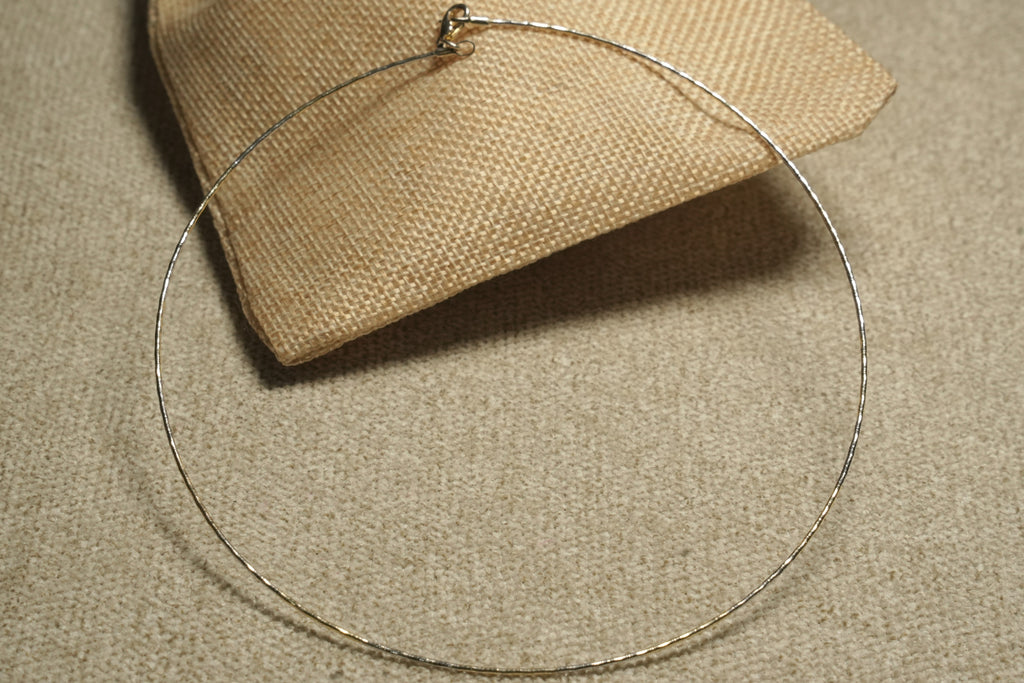 Slim gold-tone sterling silver wire collar necklace with a shimmering facet-cut surface, draped over a jute pouch.