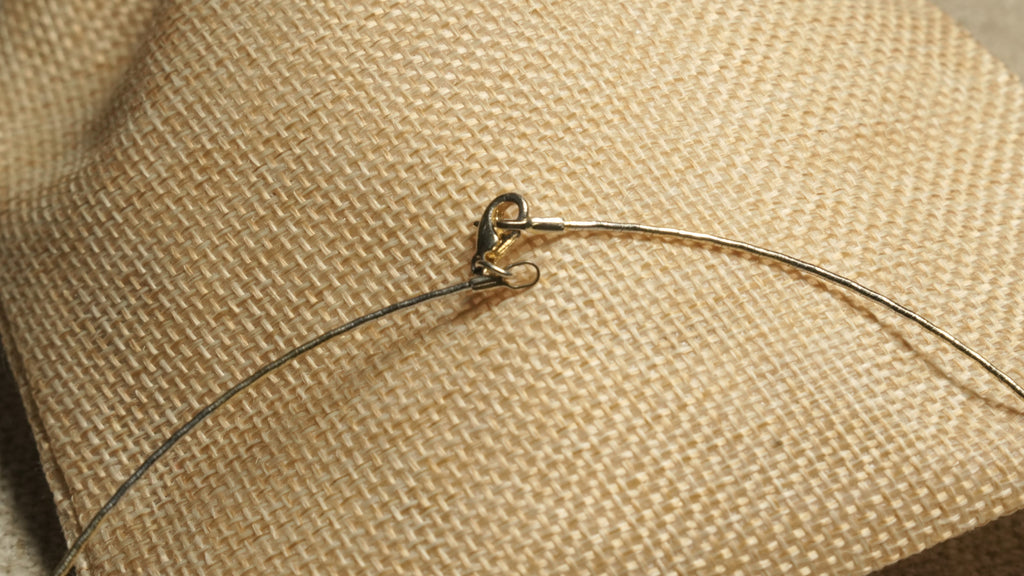Close view of the collar's lobster clasp where the gold-tone wire meets the darker grey-toned arc, resting on woven jute.