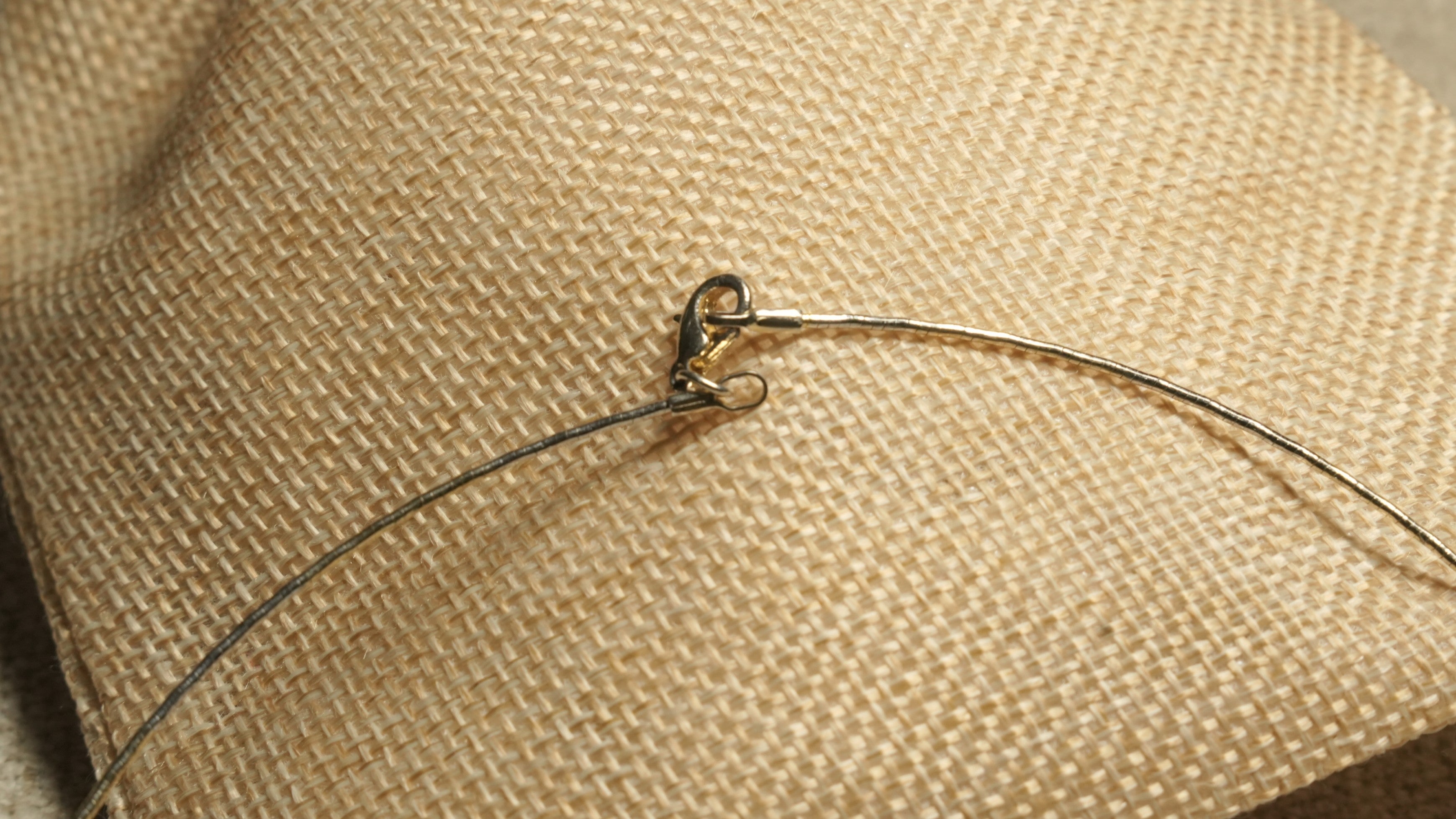 Close view of the collar's lobster clasp where the gold-tone wire meets the darker grey-toned arc, resting on woven jute.