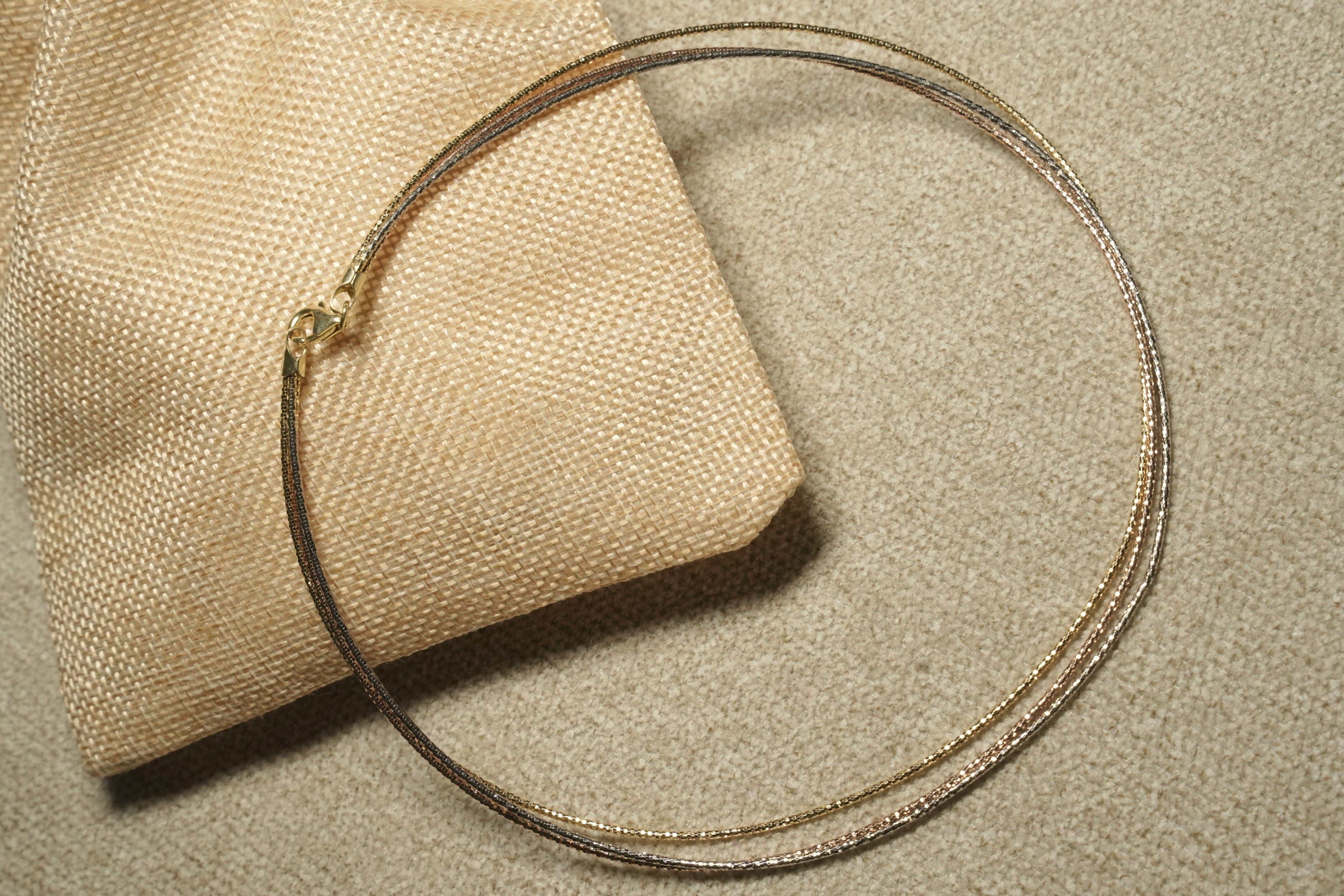 Tri-tone sterling silver wire collar necklace in gold-tone, rose gold-tone and oxidised grey, coiled over a woven jute pouch.