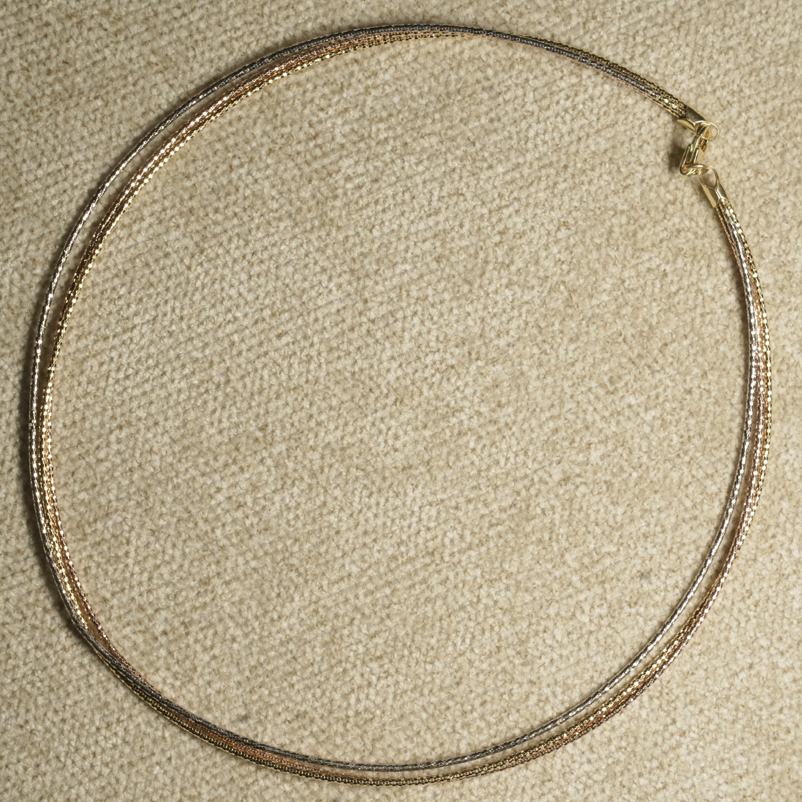 Full circle view of the three-wire collar laid flat on beige fabric, its mixed tones meeting at a gold-tone clasp.