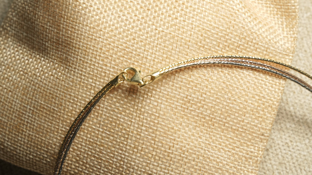 Close view of the collar's gold-tone lobster clasp and the three facet-cut wires in different tones, on woven jute.