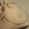 Slim gold-tone sterling silver wire collar necklace with a faceted shimmer, resting on a woven jute pouch.