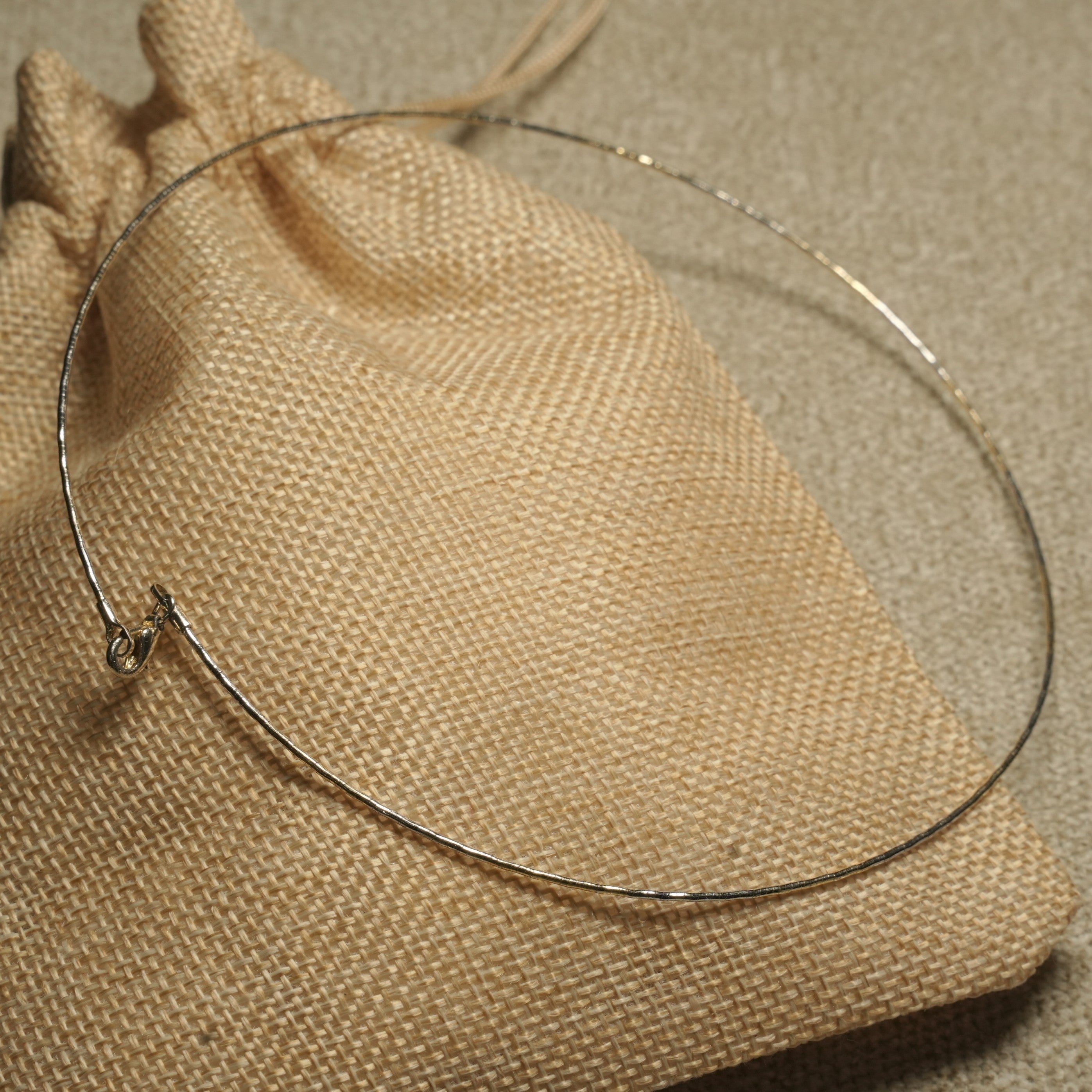 Slim gold-tone sterling silver wire collar necklace with a faceted shimmer, resting on a woven jute pouch.