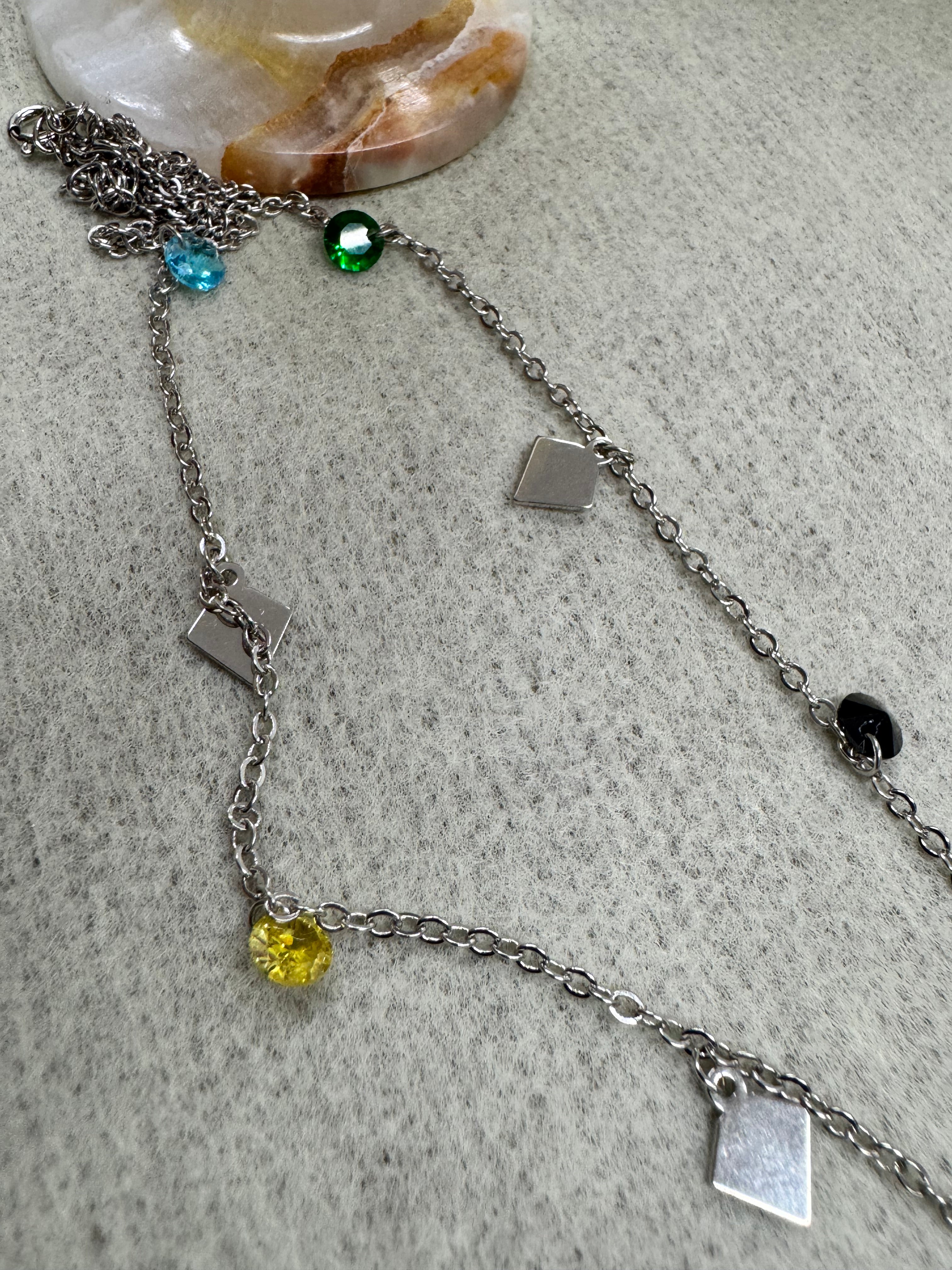 Sterling silver necklace set with mixed stones on grey felt beside a marble dish, green, blue and yellow stones spaced between kite plaques.