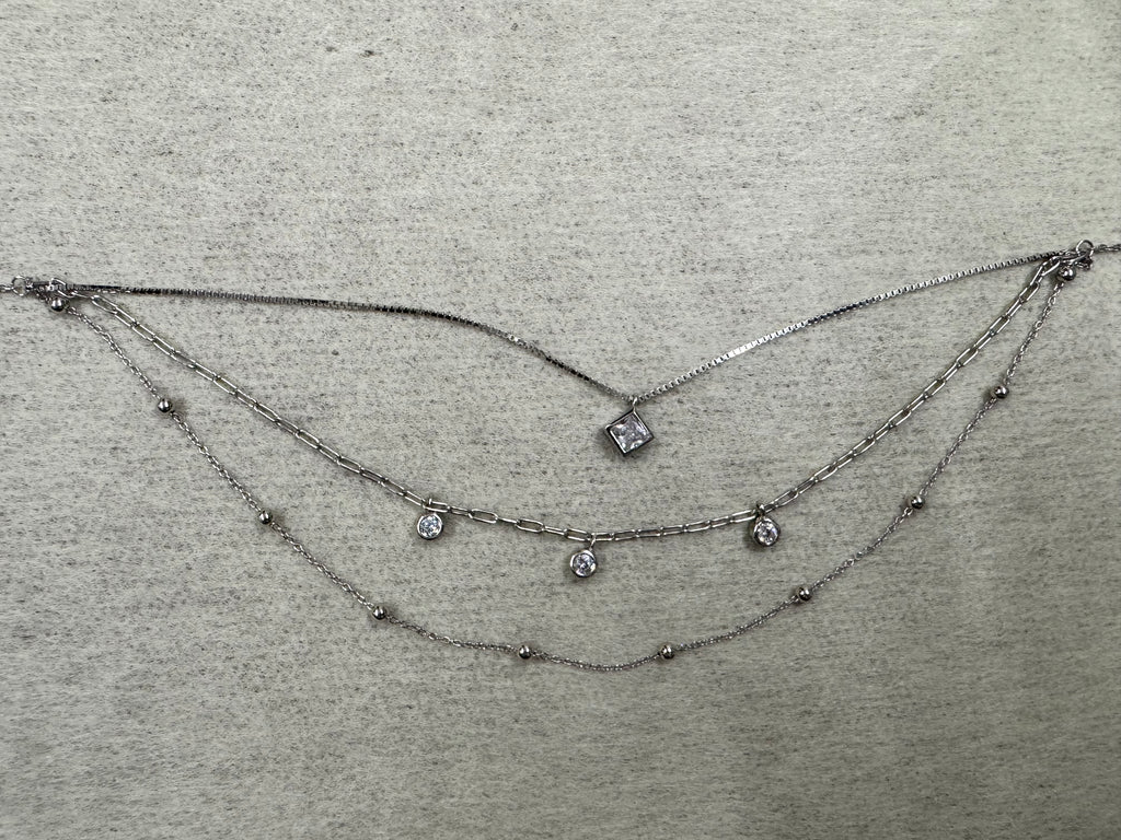 Three-strand sterling silver layered necklace: box chain with a square cubic zirconia, paperclip chain with three round cubic zirconia drops, and a satellite bead chain, laid flat on grey fabric.