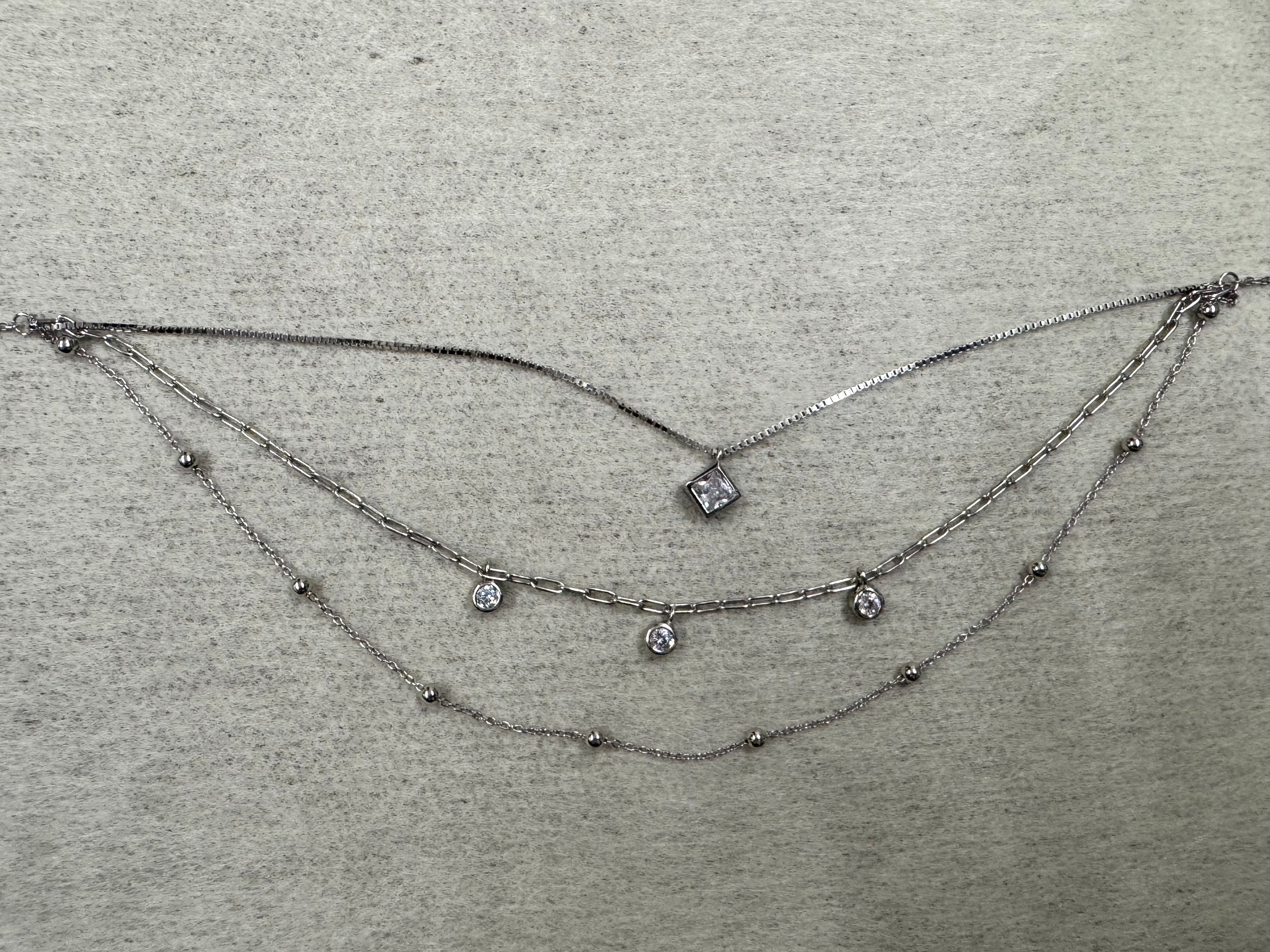 Three-strand sterling silver layered necklace: box chain with a square cubic zirconia, paperclip chain with three round cubic zirconia drops, and a satellite bead chain, laid flat on grey fabric.