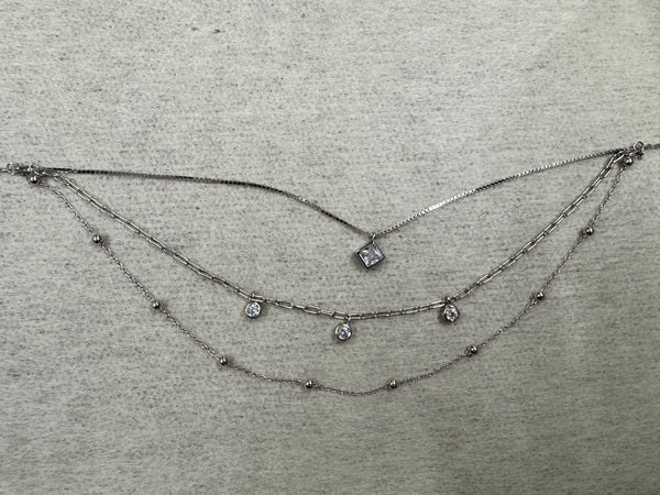 Three-strand sterling silver layered necklace: box chain with a square cubic zirconia, paperclip chain with three round cubic zirconia drops, and a satellite bead chain, laid flat on grey fabric.