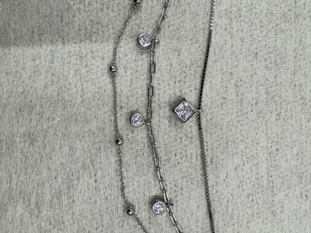 Detail of the three strands of the sterling silver necklace running side by side on grey fabric, with the square cubic zirconia on the box chain and two round stones on the paperclip chain.