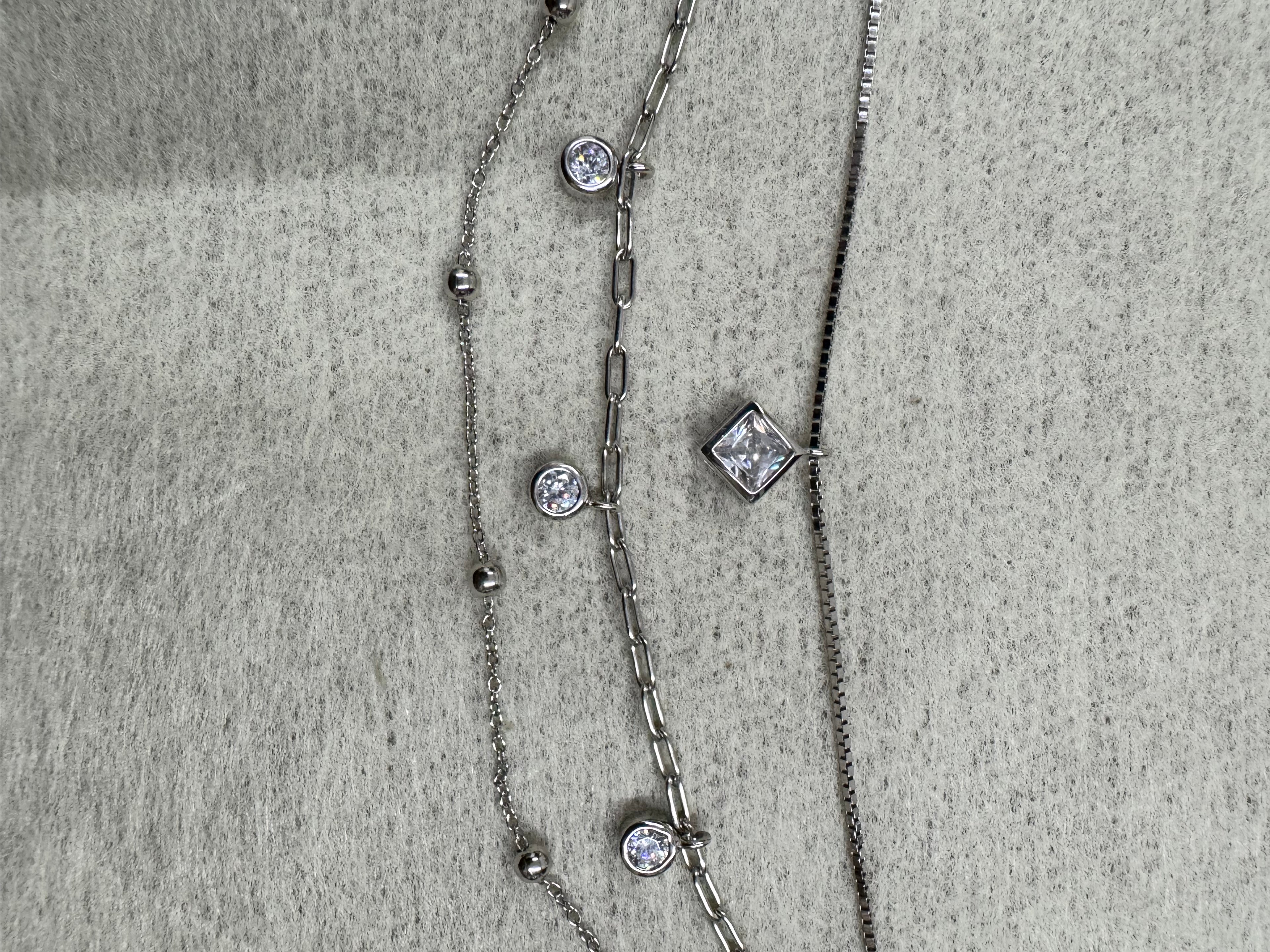 Detail of the three strands of the sterling silver necklace running side by side on grey fabric, with the square cubic zirconia on the box chain and two round stones on the paperclip chain.