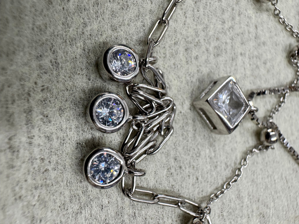 Macro view of the three round bezel-set cubic zirconia drops on the paperclip chain of the sterling silver necklace, with the square bezel to the left on grey fabric.