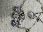 Macro view of the three round bezel-set cubic zirconia drops on the paperclip chain of the sterling silver necklace, with the square bezel to the left on grey fabric.