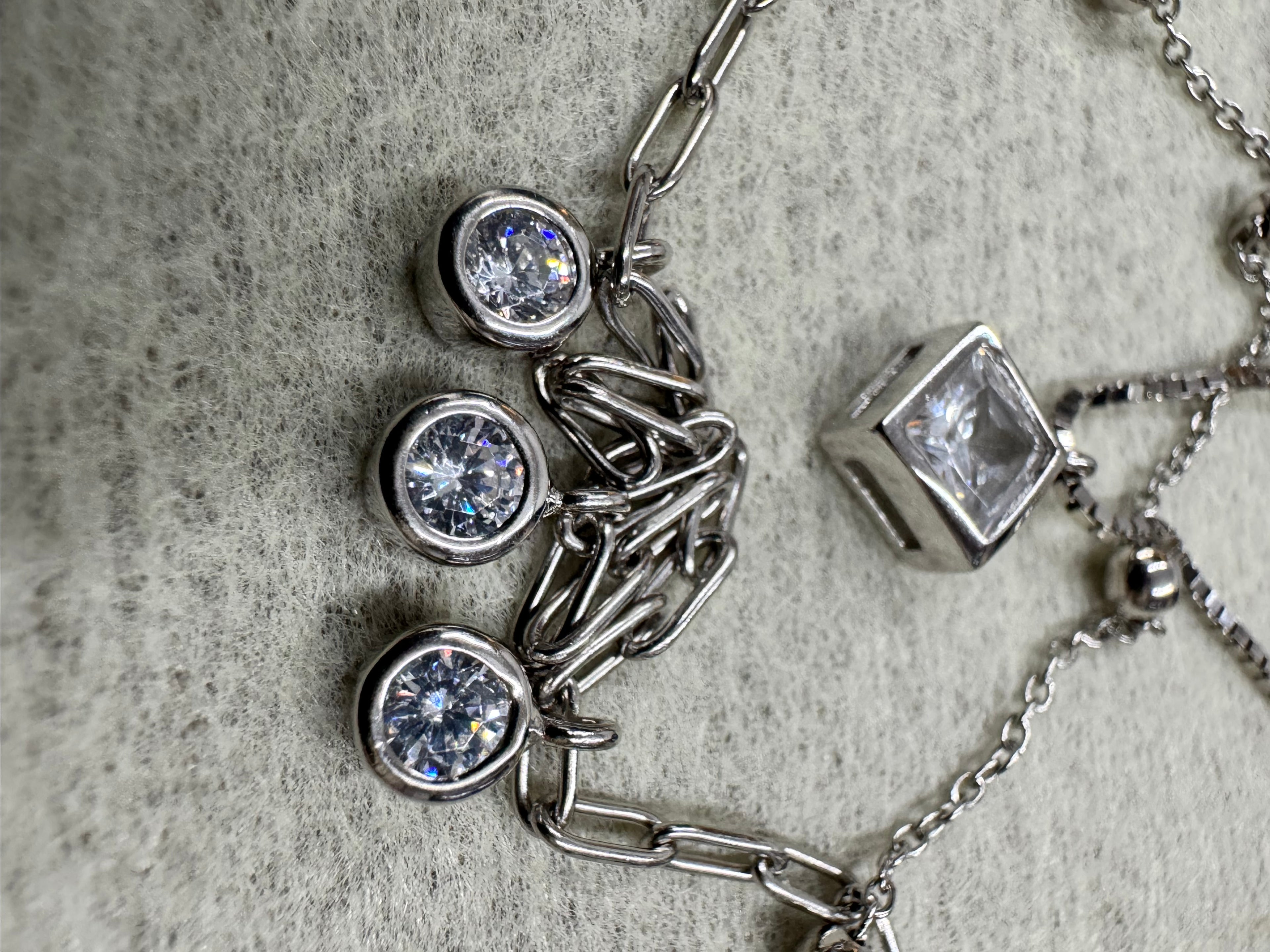 Macro view of the three round bezel-set cubic zirconia drops on the paperclip chain of the sterling silver necklace, with the square bezel to the left on grey fabric.