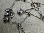 Overhead detail of the sterling silver layered necklace on grey fabric, showing the square cubic zirconia, the three round drops, the spring ring clasp and the Javeri House tag together.