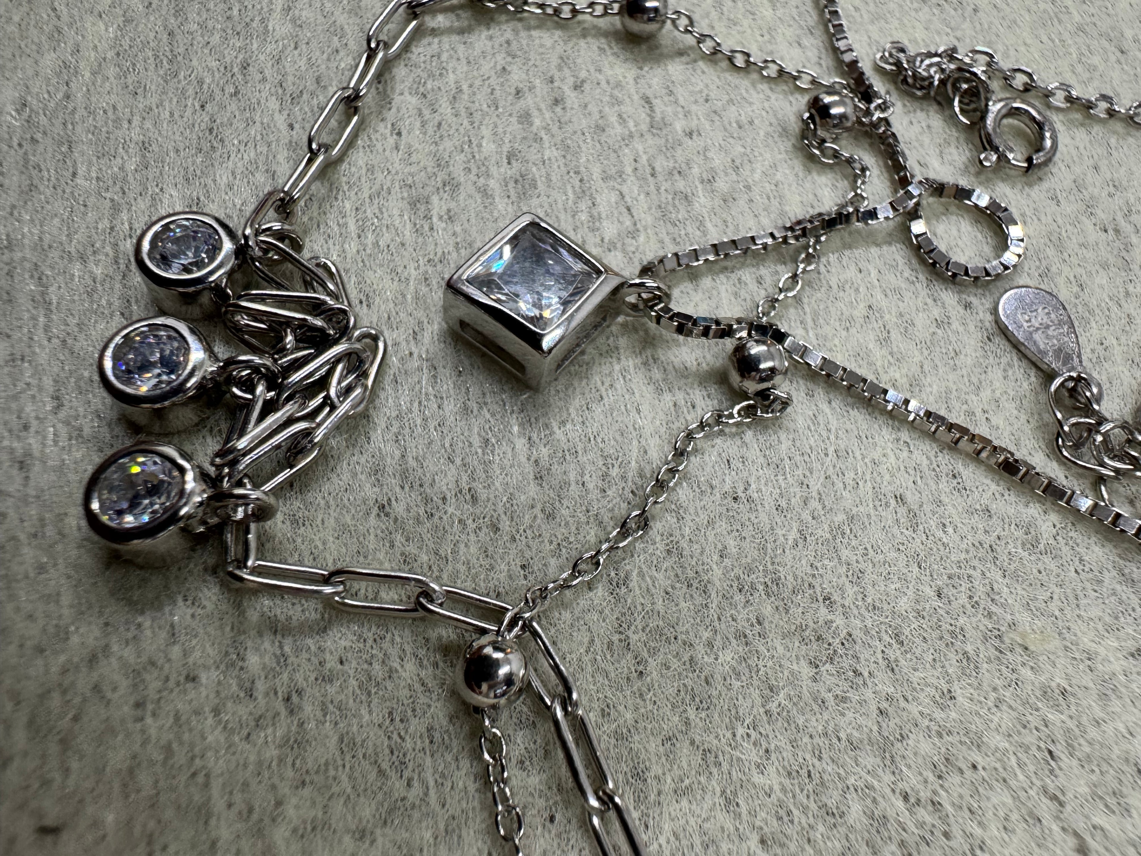 Overhead detail of the sterling silver layered necklace on grey fabric, showing the square cubic zirconia, the three round drops, the spring ring clasp and the Javeri House tag together.