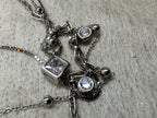 Angled close view of the sterling silver necklace with the strands crossing over one another, the square cubic zirconia and a round bezel-set stone in focus on grey fabric.