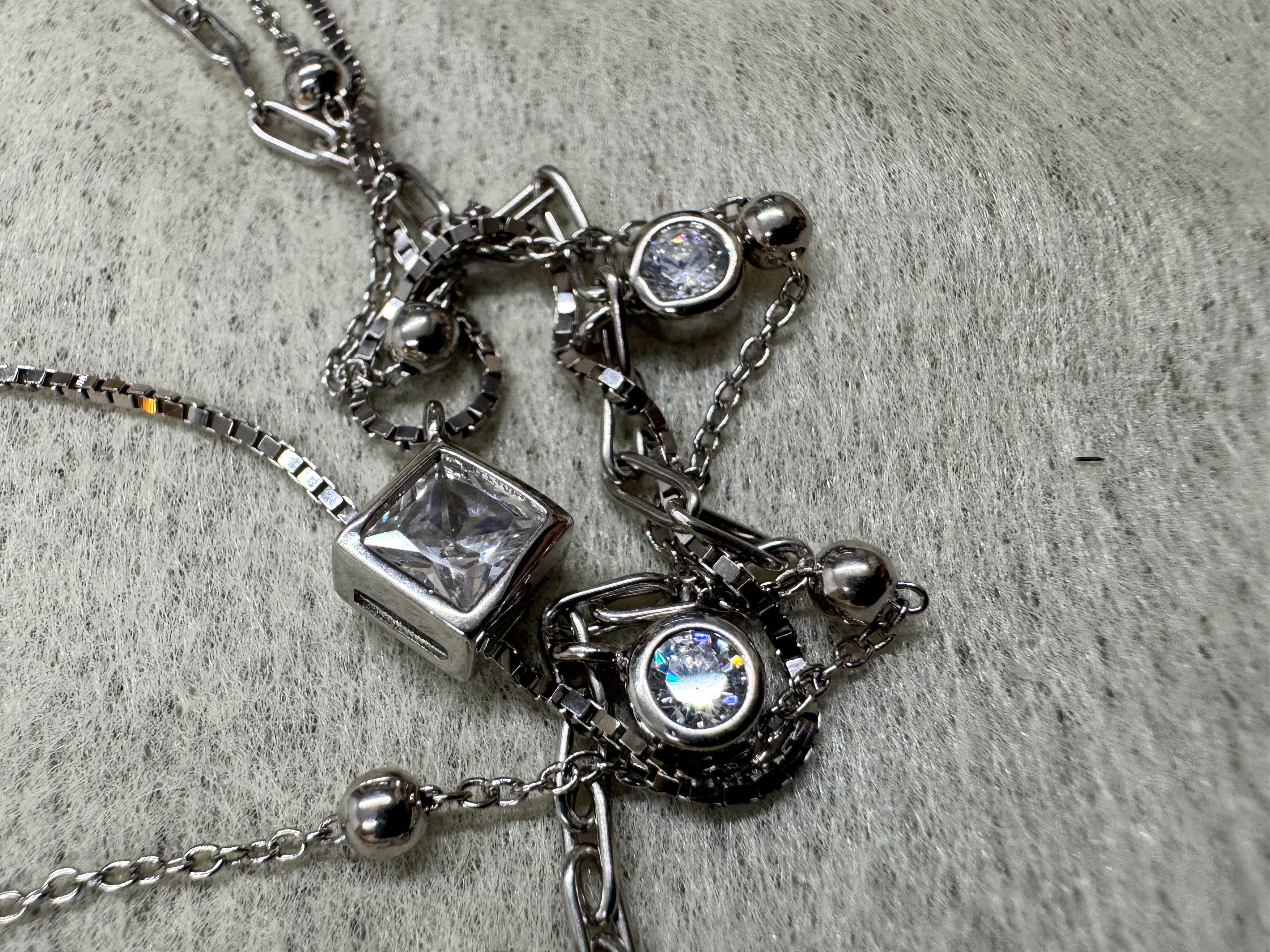 Angled close view of the sterling silver necklace with the strands crossing over one another, the square cubic zirconia and a round bezel-set stone in focus on grey fabric.
