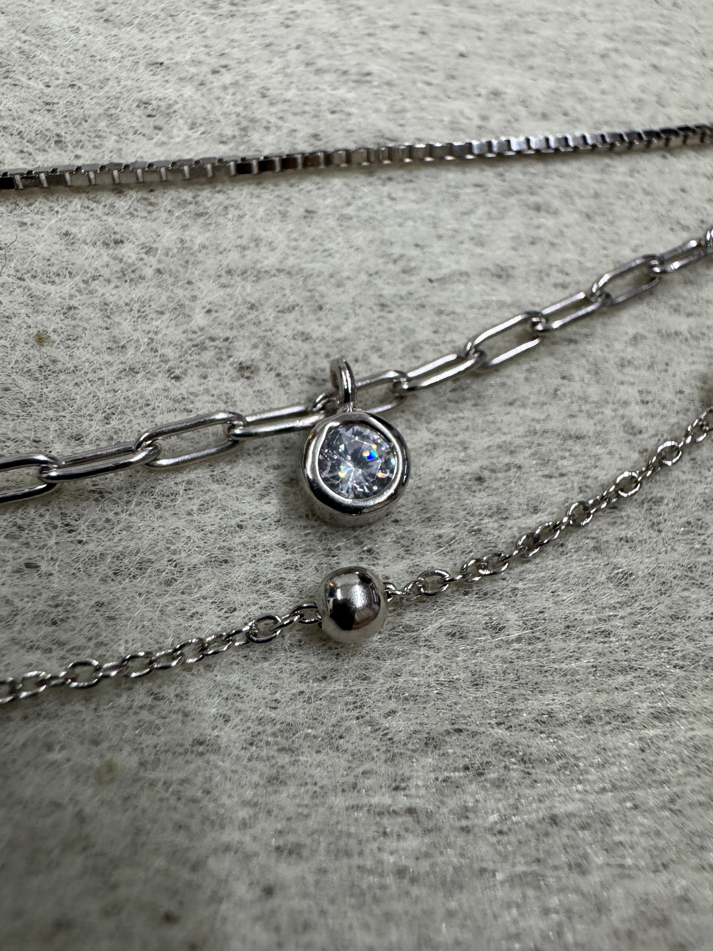Close view of the three chain constructions of the sterling silver necklace on grey fabric: box chain, paperclip chain with a round cubic zirconia and satellite chain with a polished silver ball.