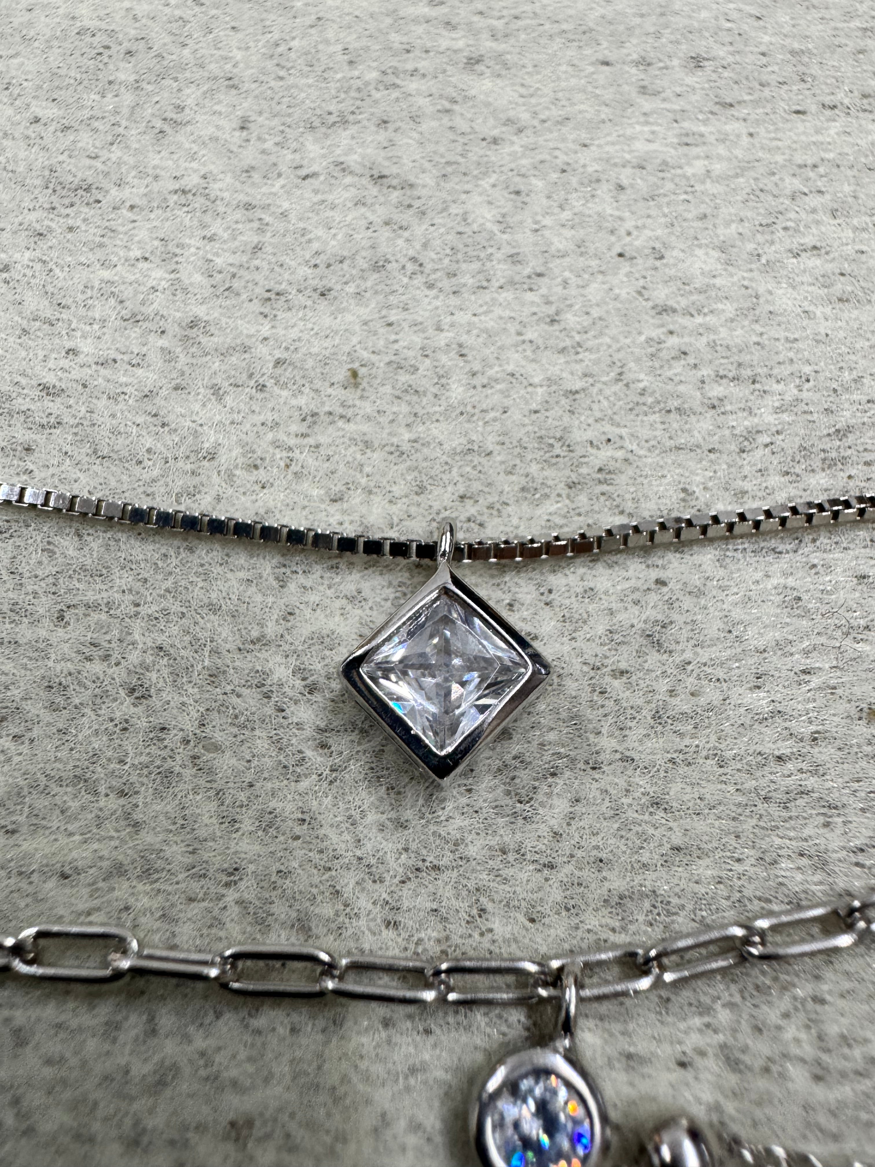 Macro of the square bezel-set cubic zirconia sitting on the fine box chain of the sterling silver necklace, the paperclip strand and a round stone at the right edge.